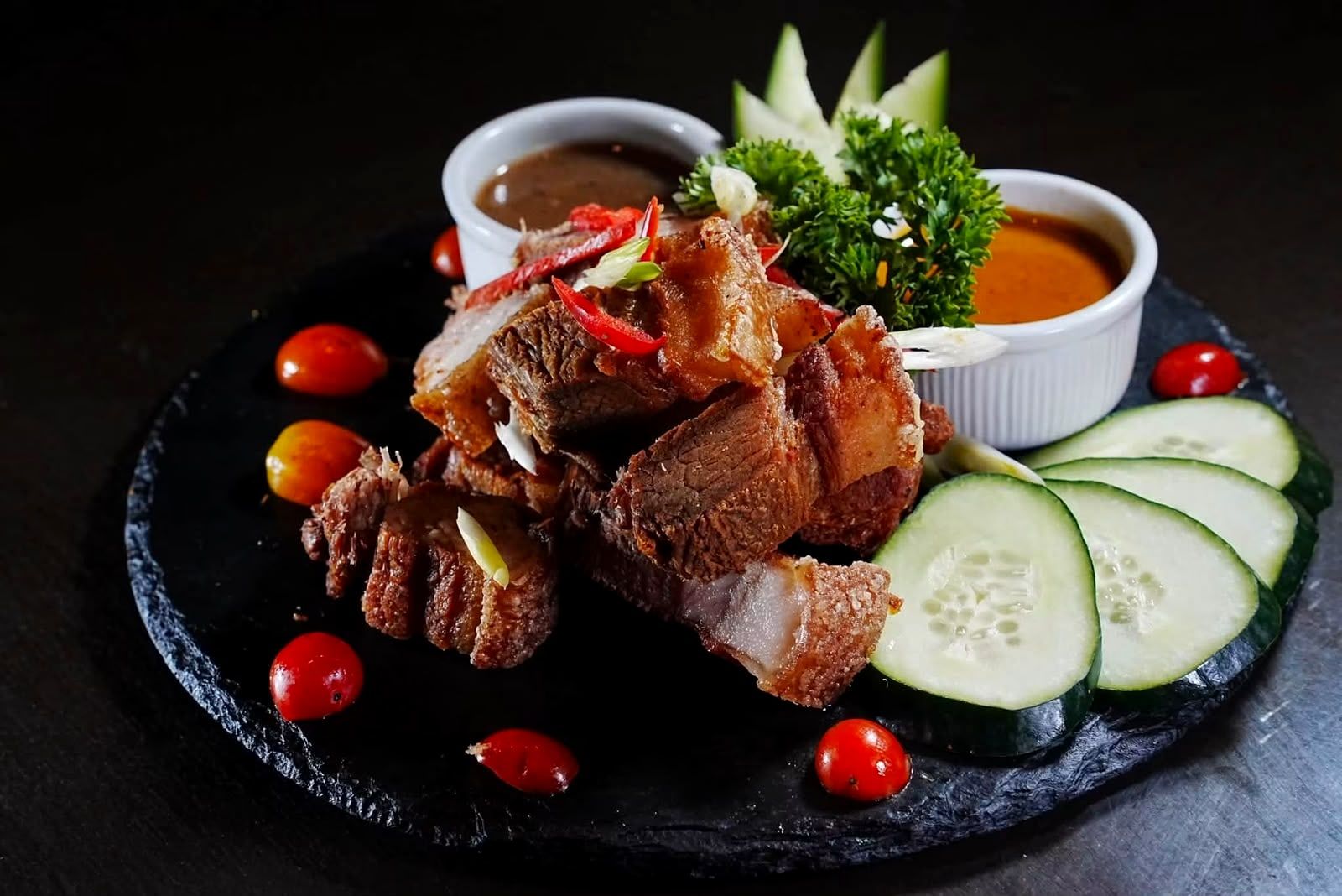 Crispy pork belly with two dipping sauces, cherry tomatoes and cucumber on a black stone plate