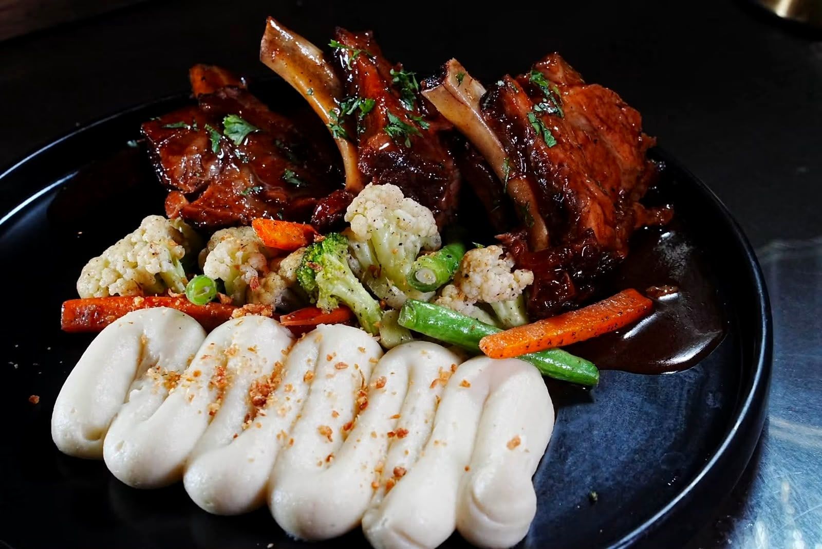 Glazed chops on the bone on a black plate with mashed potato and mixed vegetables