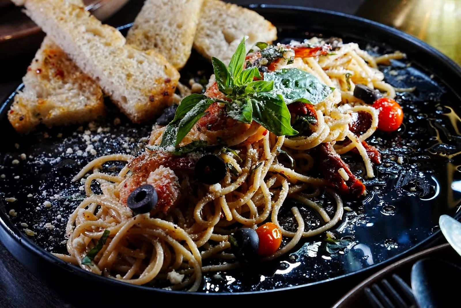 Spaghetti with olives, tomatoes and basil on a black plate with slices of toasted bread