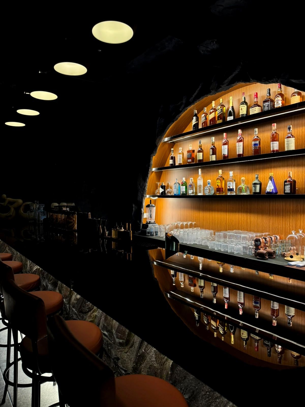 The bar at night: bottle shelves glowing inside the stone arch, with stools along the counter