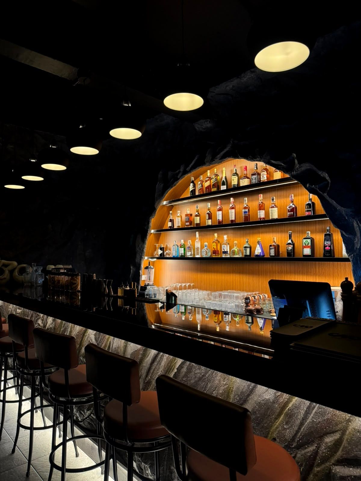 Orange bar stools along the stone faced counter under the glowing arched shelves