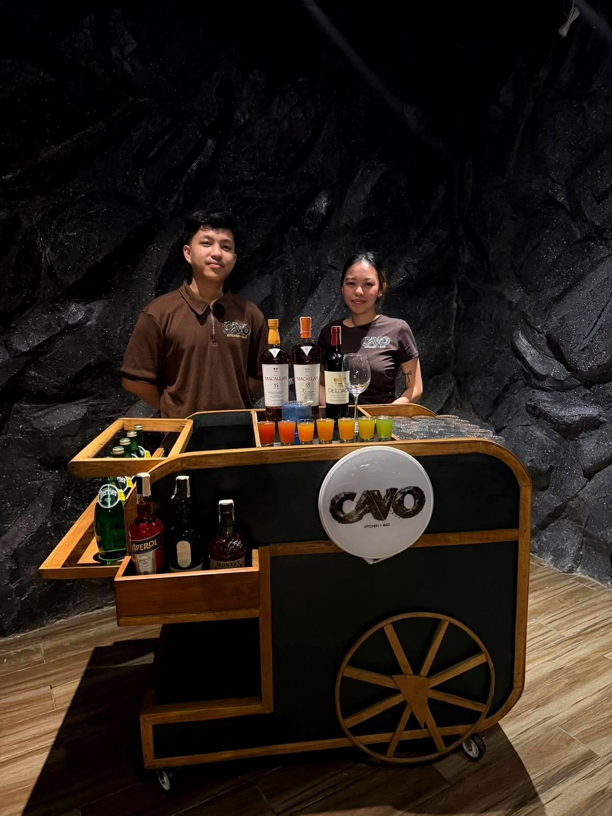 Two Cavo staff in brown uniforms standing behind the Cavo mobile bar, in front of a rock wall