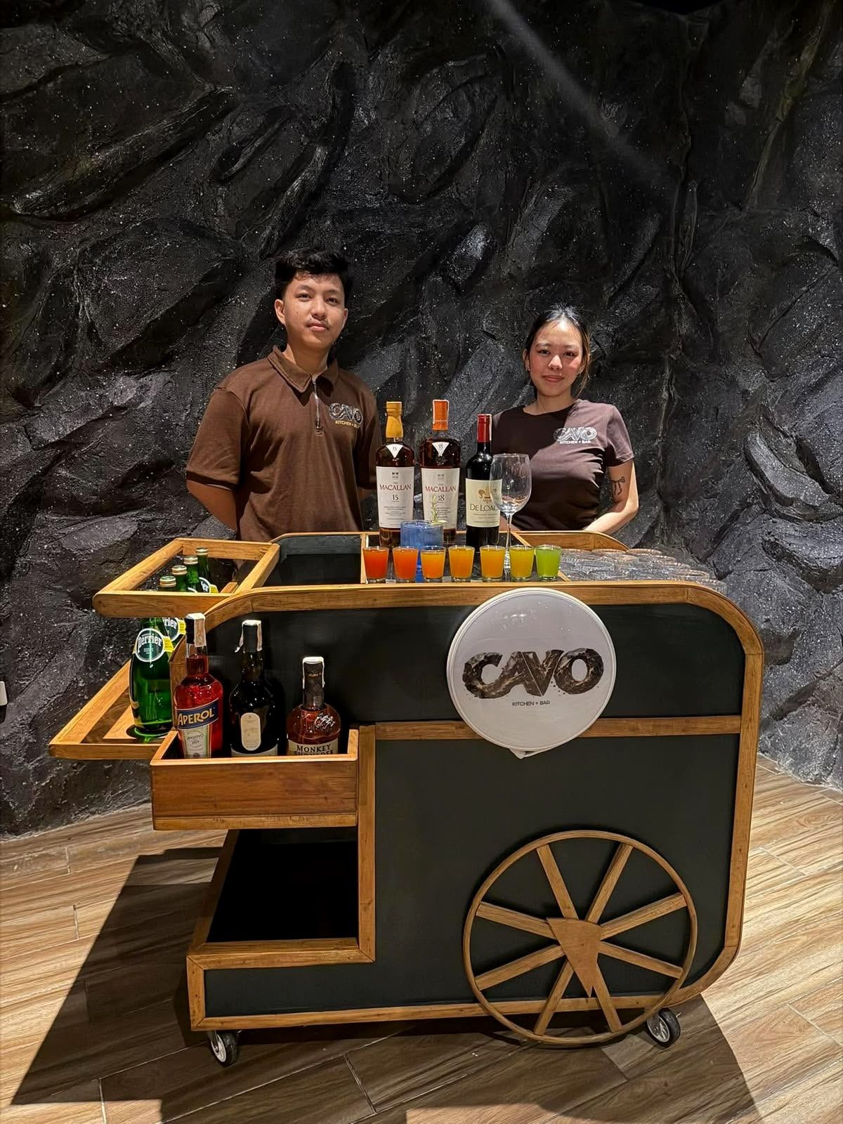 Two Cavo staff standing behind the wooden mobile bar cart with bottles and shots