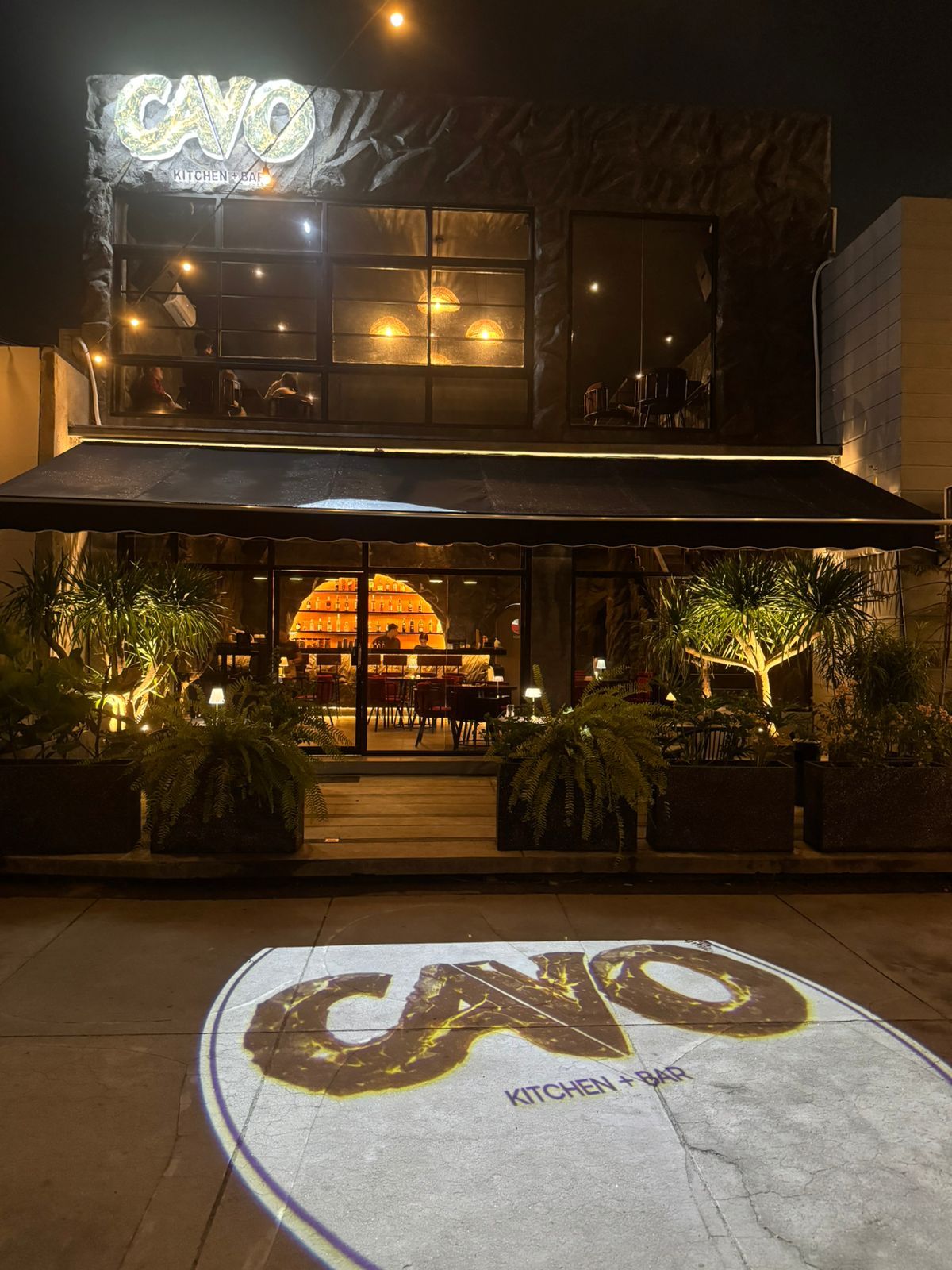 The front of Cavo at night: the lit CAVO sign above the glass entrance and the logo projected onto the pavement