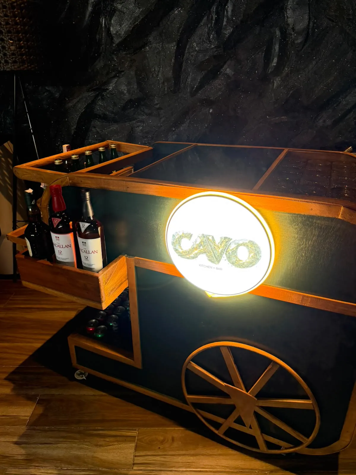 The Cavo mobile bar cart with the lit CAVO logo disc, bottles on the shelf and a wooden wheel, in front of a rock wall