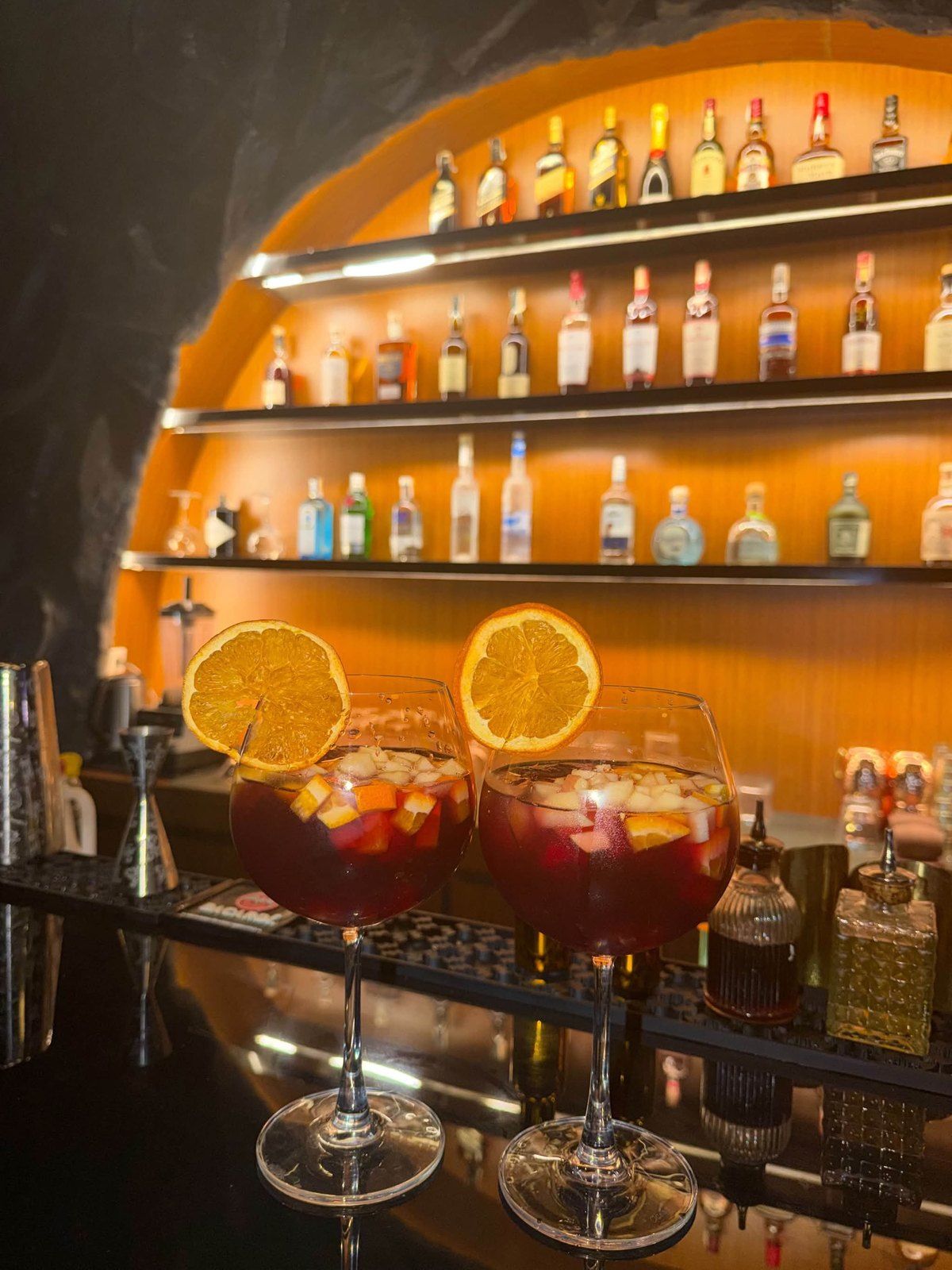 Two glasses of sangria with orange slices on the bar, under the lit bottle shelves