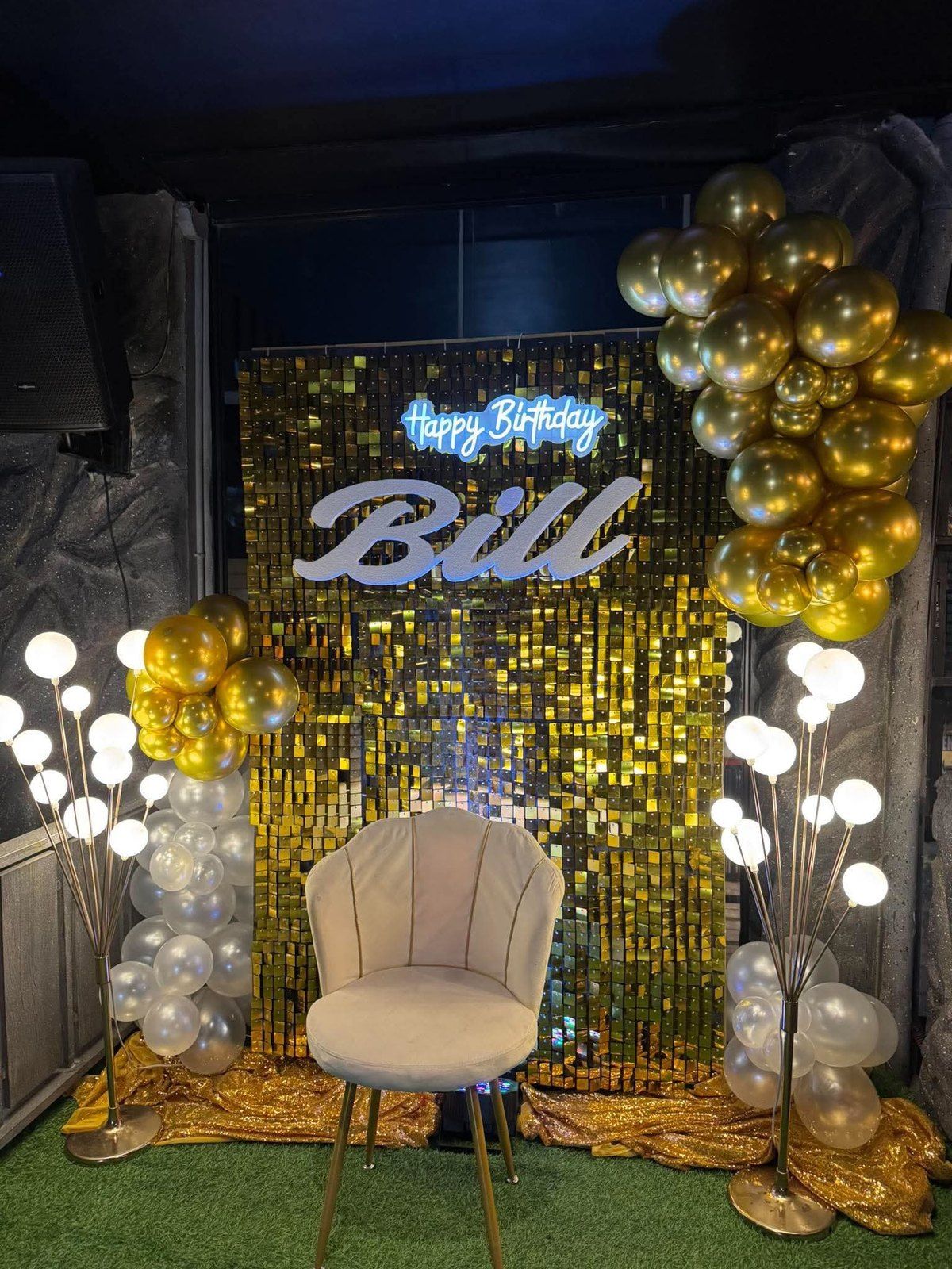 A birthday setup at Cavo: a gold sequin wall with a lit name sign, gold and silver balloons and glowing lamps