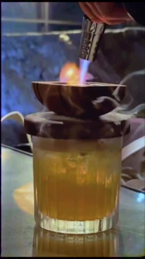 A torch lights the smoker on a Sats Cask, a cask lid traps the smoke, then the glass is opened