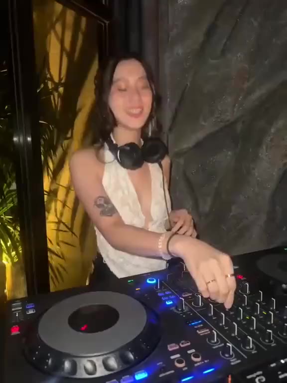 A DJ in a white lace top, headphones on, smiling at the decks