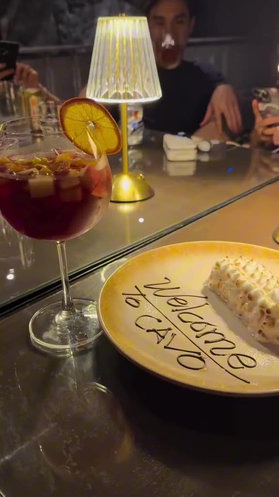 A Welcome to Cavo dessert plate and a sangria with an orange slice under a table lamp